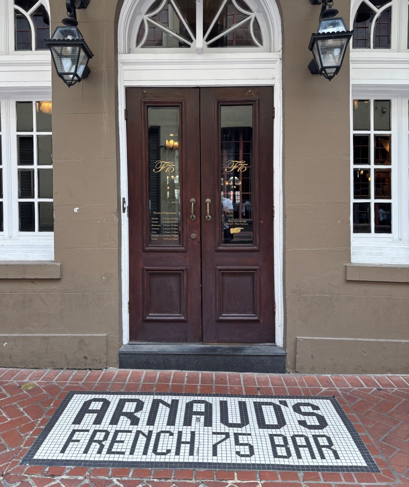 Entrance to Arnaud's French 75 Bar with double wooden doors and mosaic tile sign on the ground.