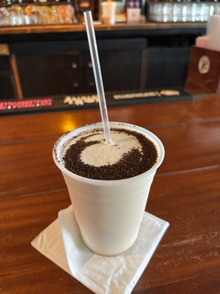 Creamy drink in a white cup with a straw, sprinkled with chocolate powder, on a wooden table.